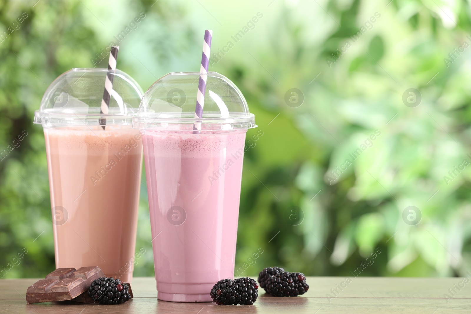 Photo of Tasty milkshakes in plastic cups and ingredients on wooden table outdoors, space for text