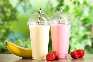 Photo of Tasty milkshakes in plastic cups and ingredients on wooden table outdoors