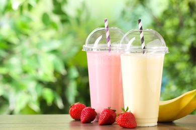 Photo of Tasty milkshakes in plastic cups and ingredients on wooden table outdoors, space for text