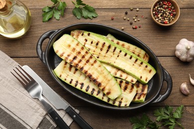 Photo of Delicious grilled courgette slices in baking dish, oil and spices on wooden table, flat lay