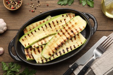 Photo of Delicious grilled courgette slices in baking dish, cutlery and spices on wooden table, flat lay