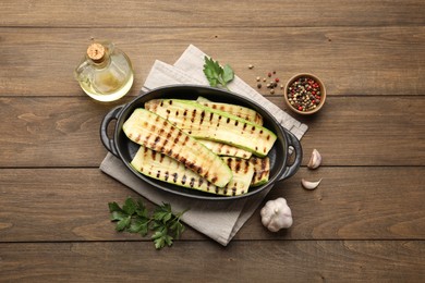 Photo of Delicious grilled courgette slices in baking dish, oil and spices on wooden table, flat lay