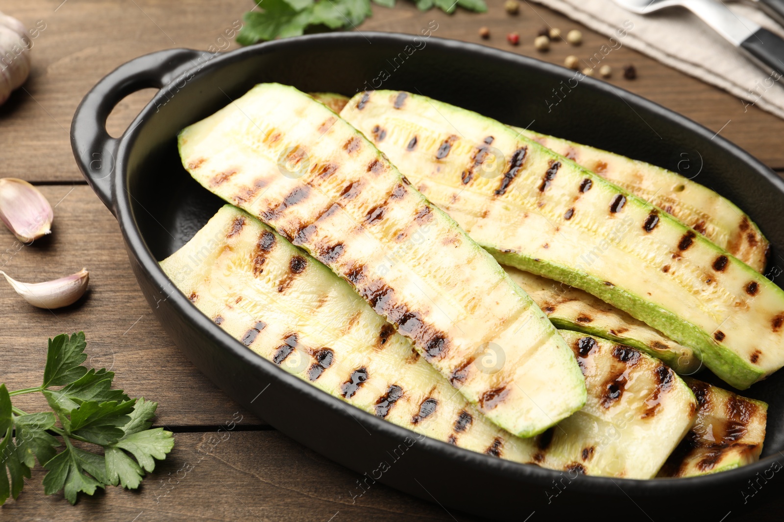 Photo of Delicious grilled courgette slices in baking dish and parsley on wooden table, closeup