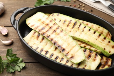 Photo of Delicious grilled courgette slices in baking dish and parsley on wooden table, closeup