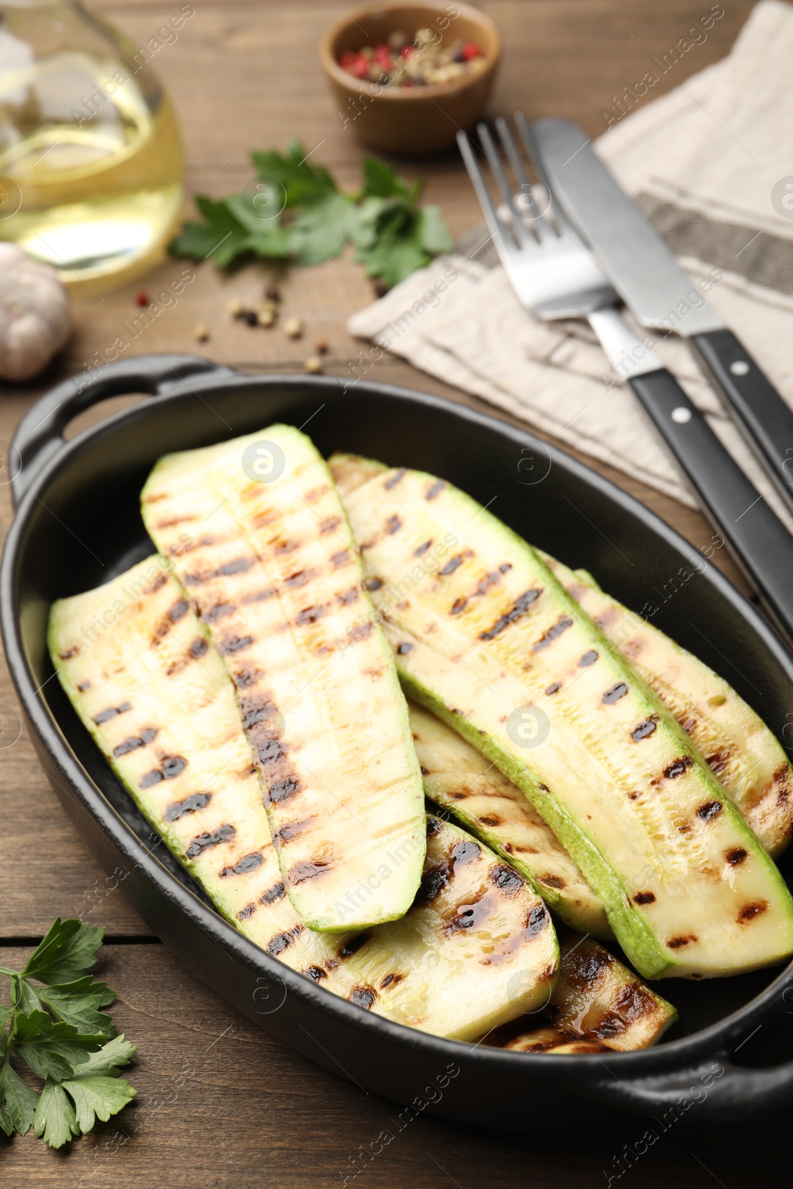 Photo of Delicious grilled courgette slices in baking dish and parsley on wooden table, closeup