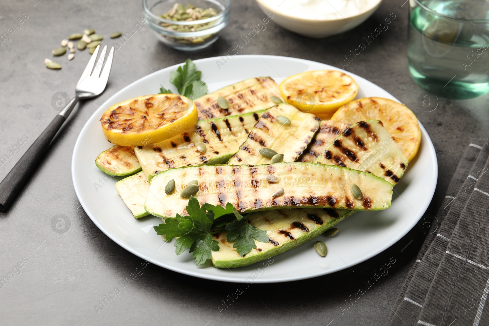 Photo of Delicious grilled courgette slices with pumpkin seeds, lemon and parsley on grey table, closeup