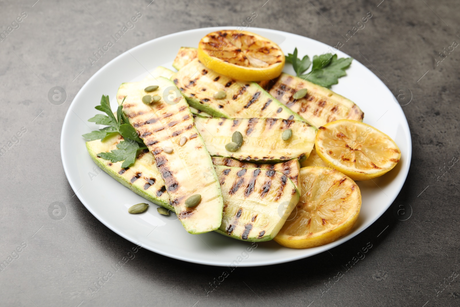 Photo of Delicious grilled courgette slices with pumpkin seeds, lemon and parsley on grey table, closeup