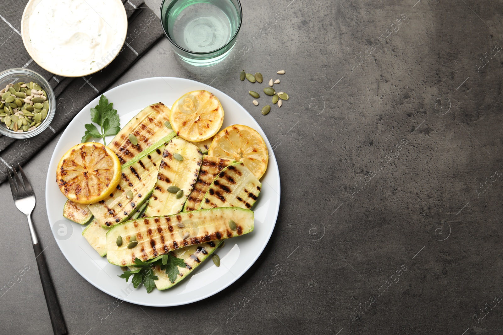 Photo of Delicious grilled courgette slices with pumpkin seeds, lemon and parsley served on grey textured table, flat lay. Space for text