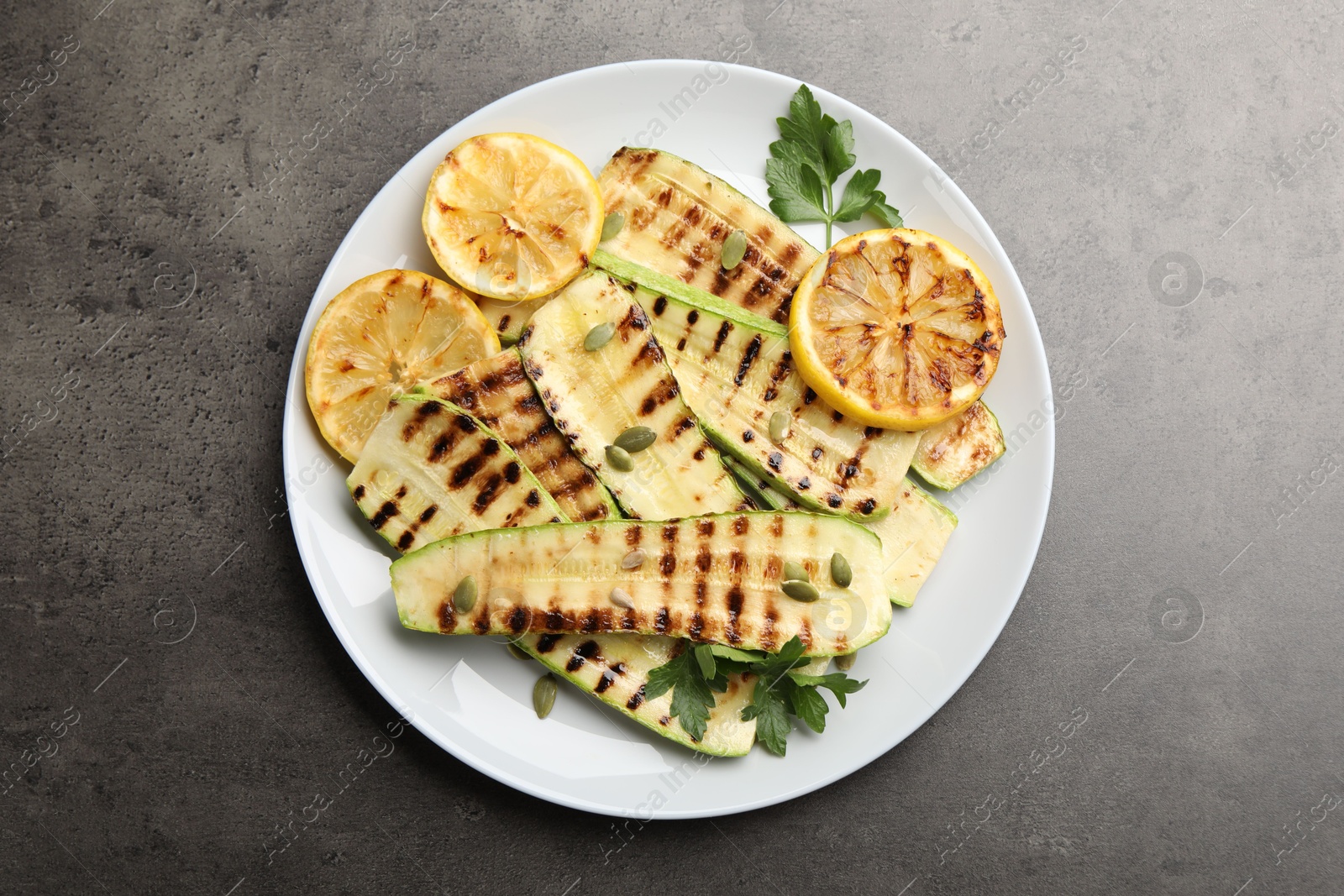 Photo of Delicious grilled courgette slices with pumpkin seeds, lemon and parsley on grey textured table, top view