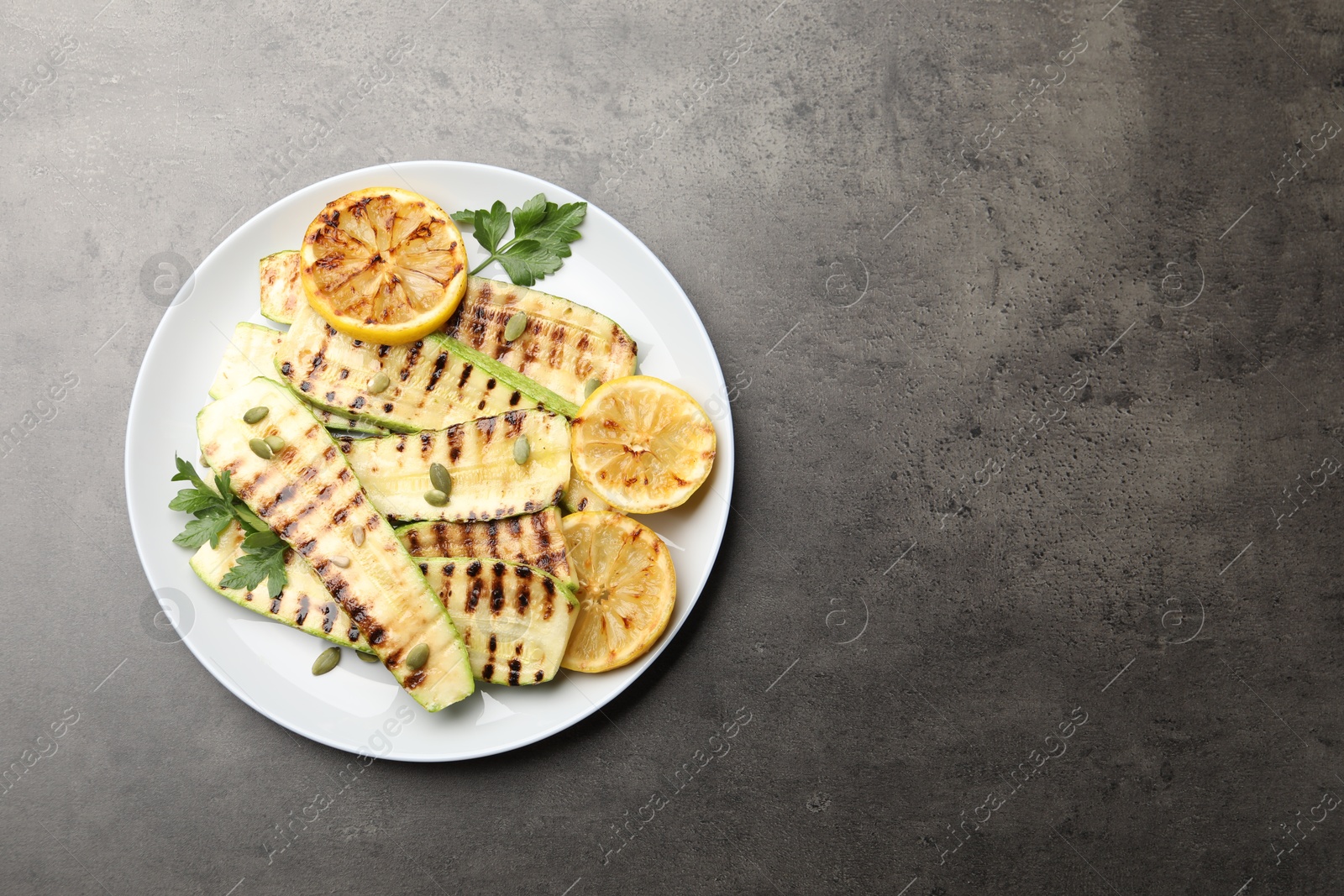 Photo of Delicious grilled courgette slices with pumpkin seeds, lemon and parsley on grey textured table, top view. Space for text