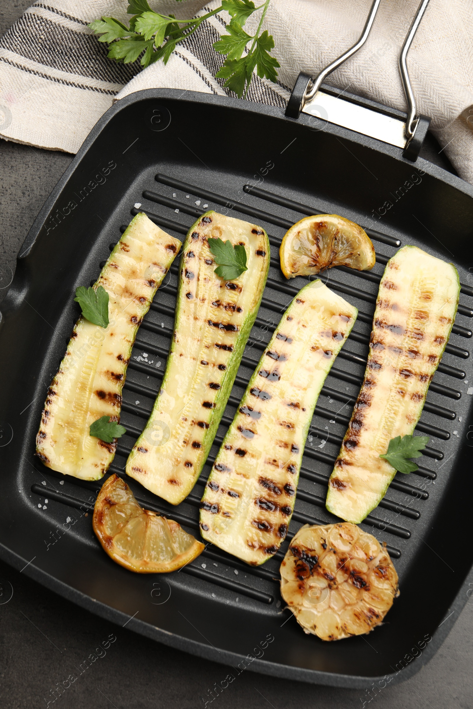 Photo of Delicious grilled courgette slices with garlic, lemon and parsley in pan on grey textured table, flat lay