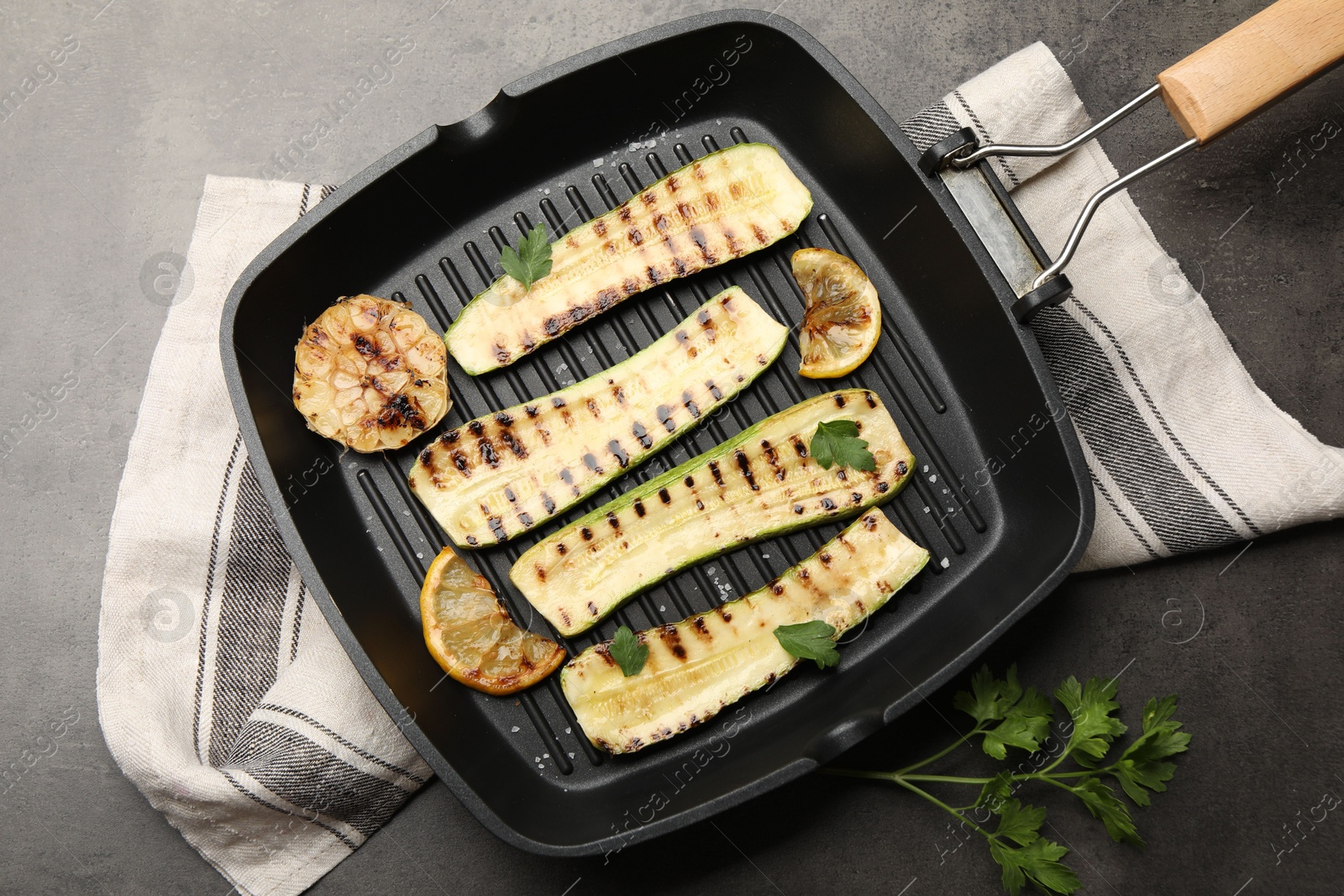Photo of Delicious grilled courgette slices with garlic, lemon and parsley in pan on grey textured table, flat lay