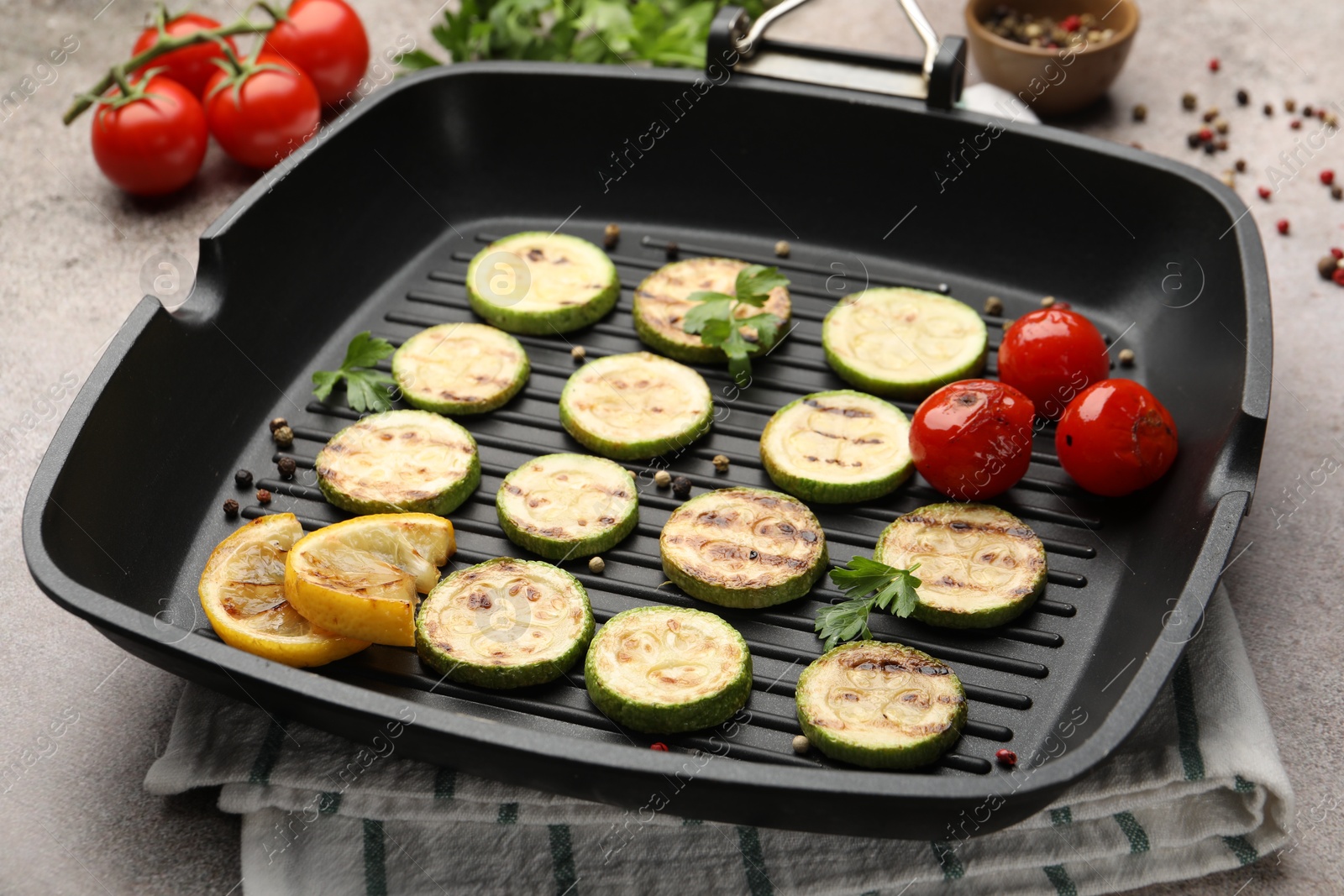 Photo of Delicious grilled courgette slices with tomatoes, lemon and parsley in pan on grey textured table, closeup