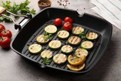 Photo of Delicious grilled courgette slices with tomatoes, lemon and parsley in pan on grey textured table, closeup