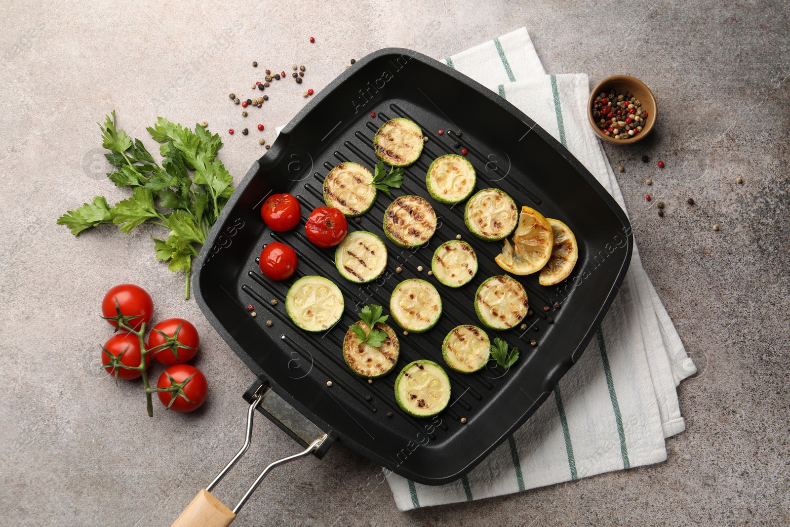 Photo of Delicious grilled courgette slices with tomatoes, lemon and parsley in pan on grey textured table, flat lay