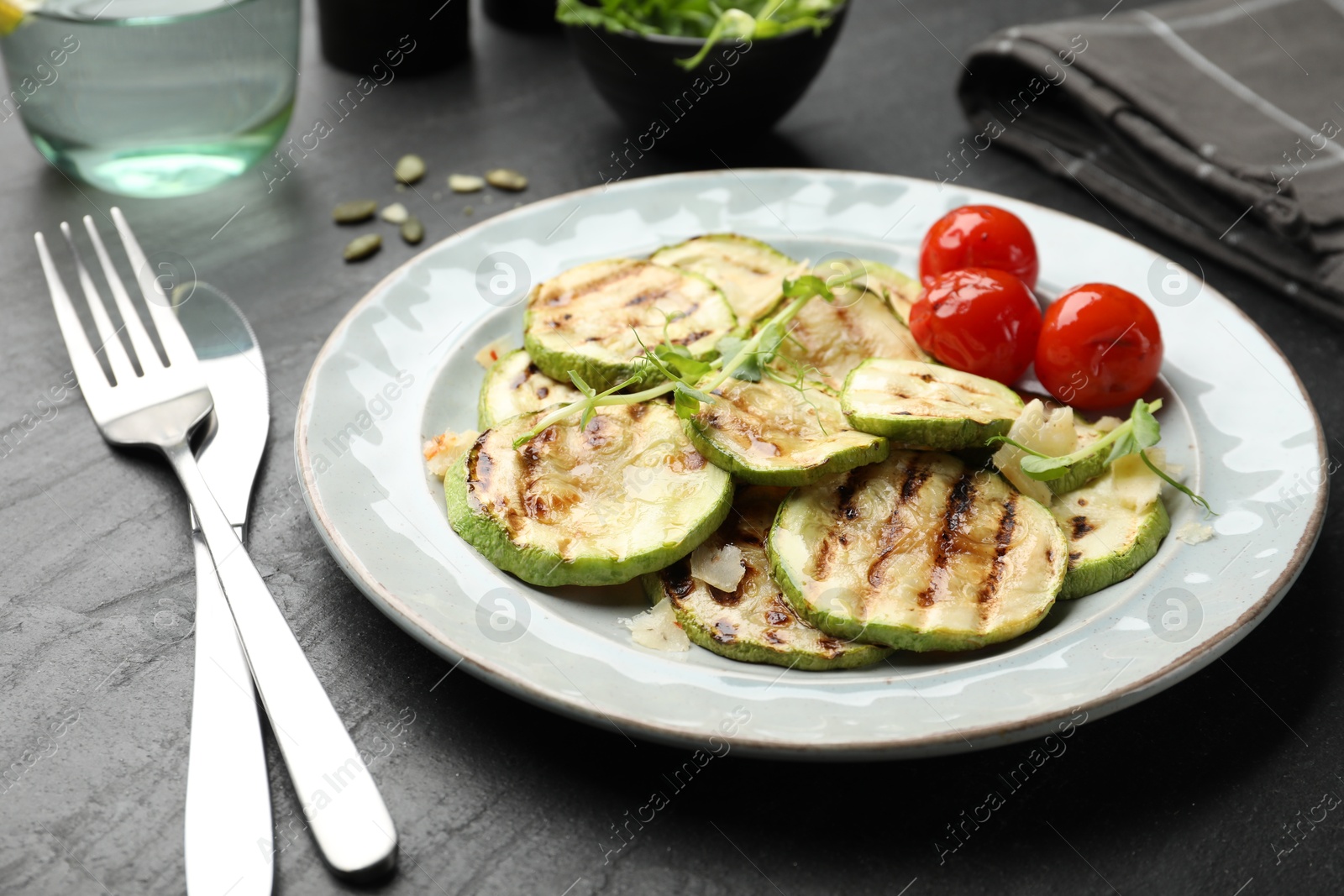 Photo of Delicious grilled courgette slices with tomatoes and microgreens served on black textured table, closeup