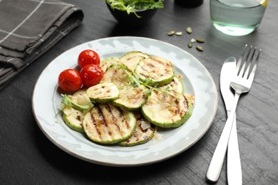 Photo of Delicious grilled courgette slices with tomatoes and microgreens served on black textured table, closeup