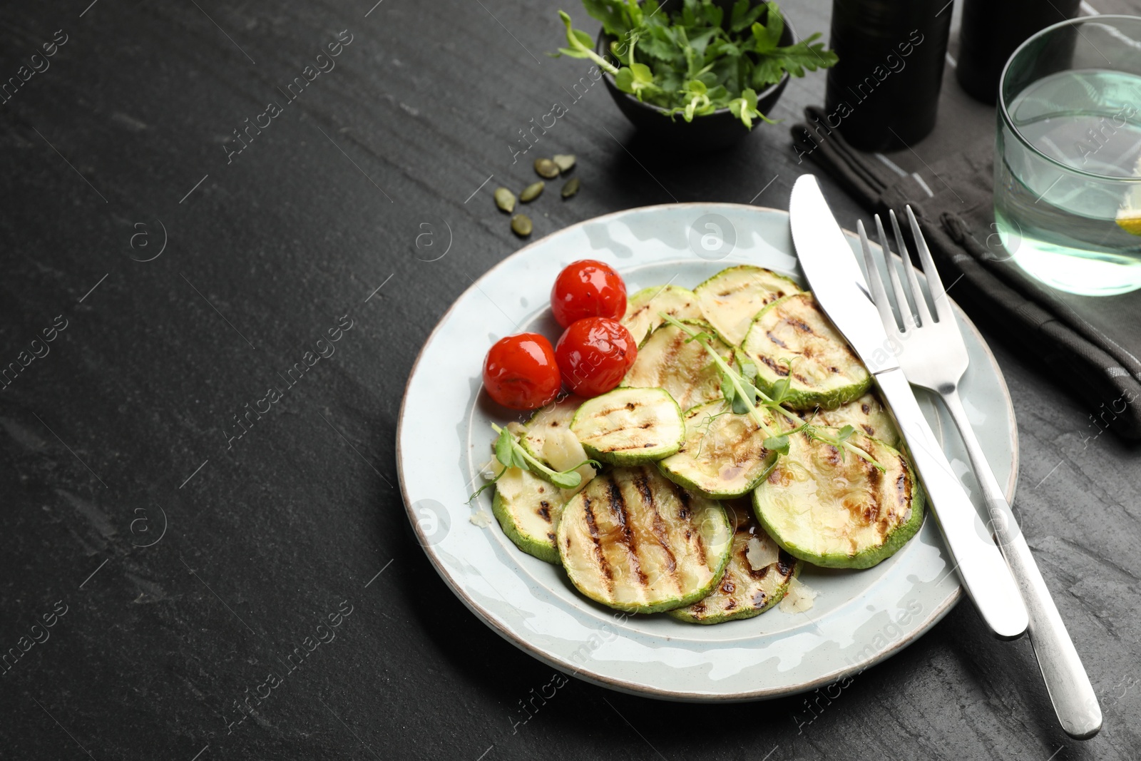 Photo of Delicious grilled courgette slices with tomatoes and microgreens served on black textured table, closeup