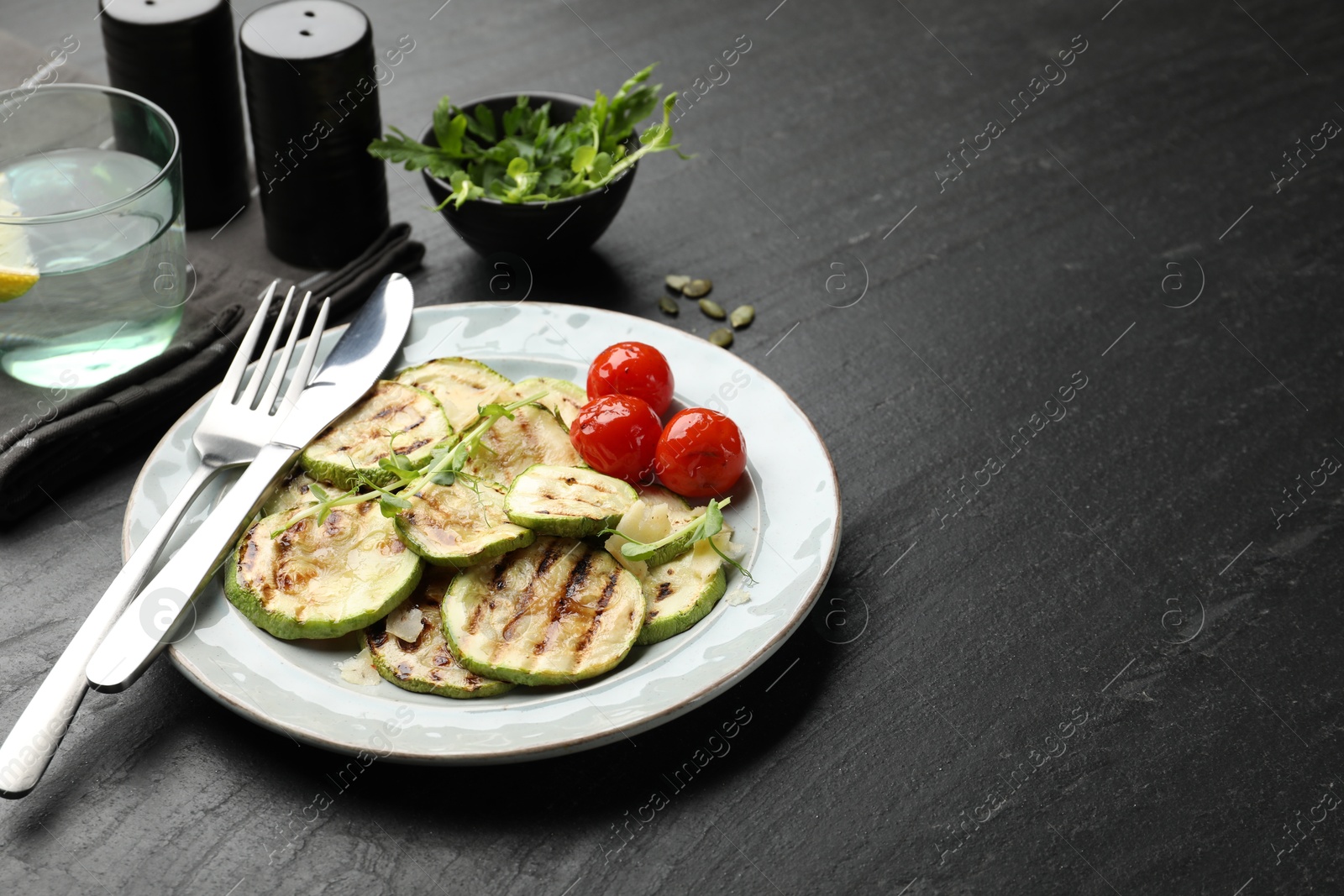 Photo of Delicious grilled courgette slices with tomatoes and microgreens served on black textured table, closeup