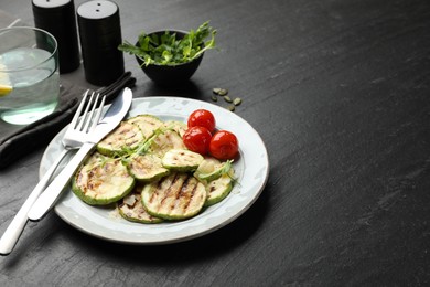 Photo of Delicious grilled courgette slices with tomatoes and microgreens served on black textured table, closeup