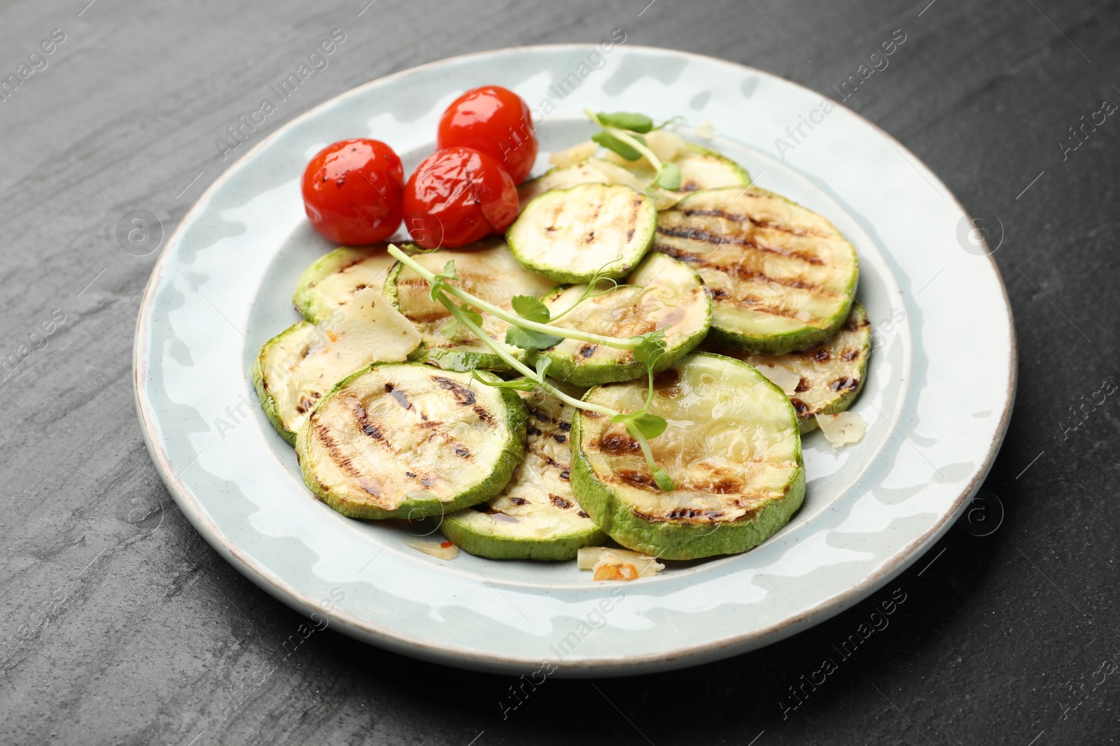 Photo of Delicious grilled courgette slices with tomatoes and microgreens on black textured table, closeup
