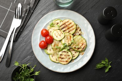 Photo of Delicious grilled courgette slices with tomatoes and microgreens served on black textured table, flat lay