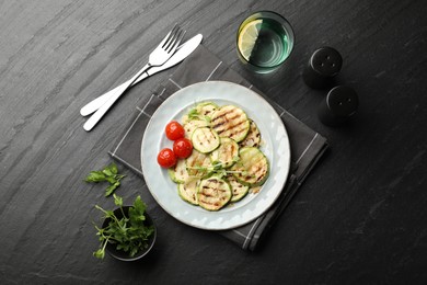 Photo of Delicious grilled courgette slices with tomatoes and microgreens served on black textured table, flat lay