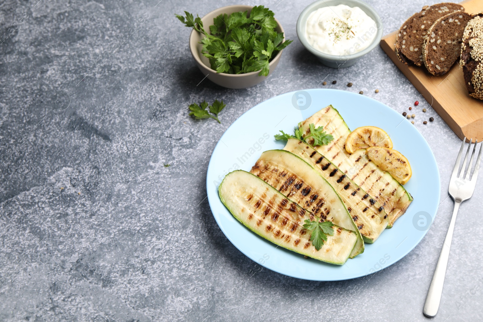 Photo of Delicious grilled courgette slices with parsley and lemon served on gray textured table. Space for text