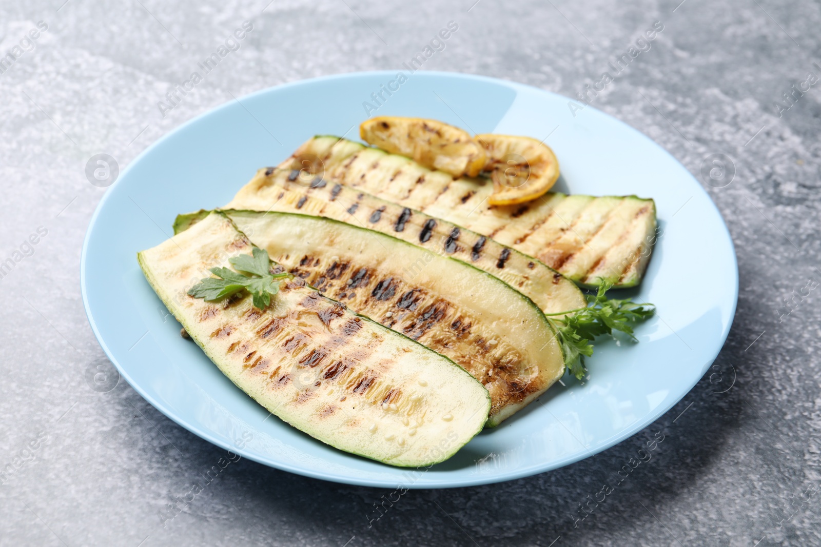 Photo of Delicious grilled courgette slices with parsley and lemon on gray textured table, closeup