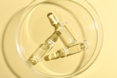 Photo of Glass ampoules with liquid and Petri dish on yellow background, top view
