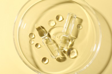 Photo of Glass ampoules with liquid and Petri dish on yellow background, top view