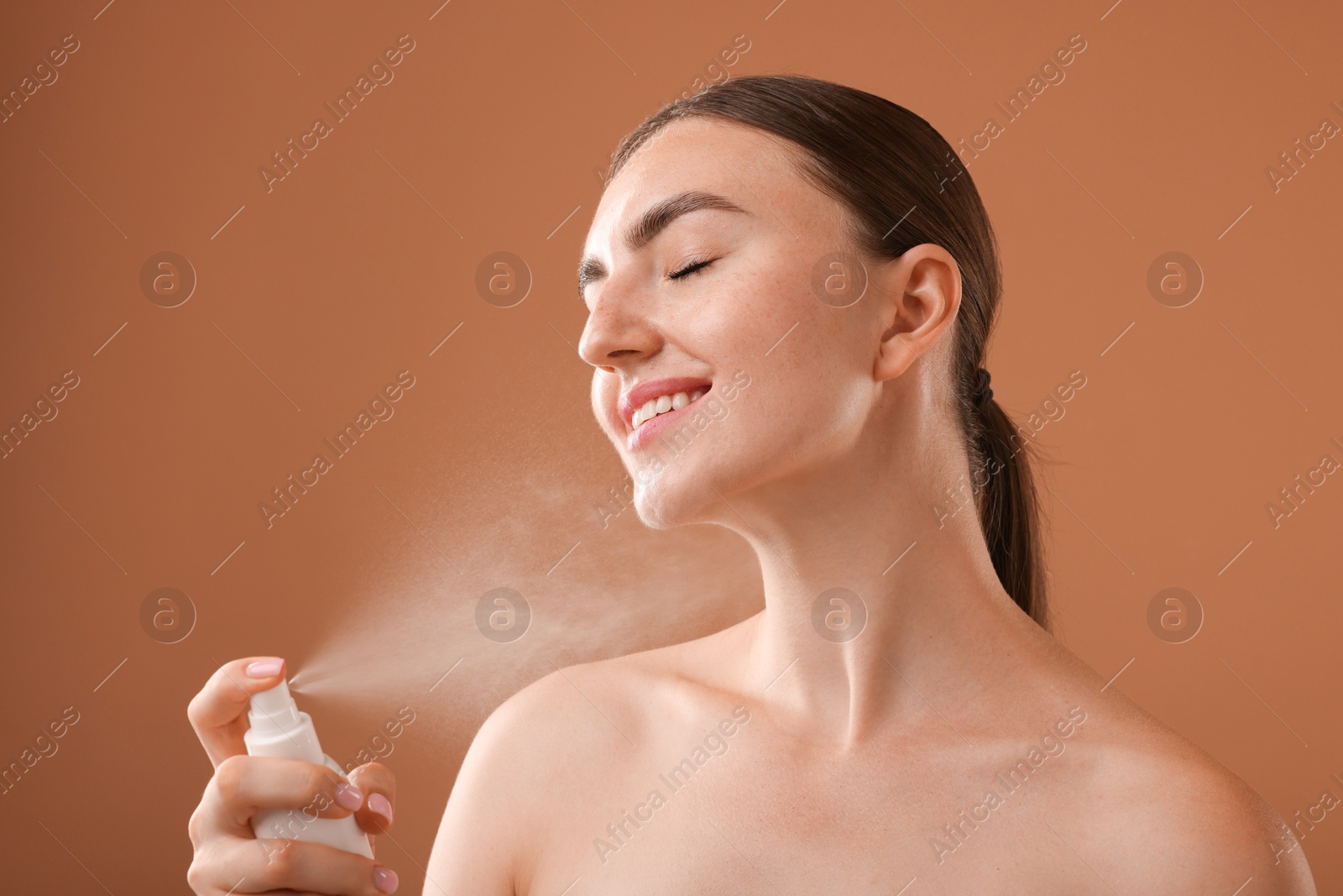 Smiling woman spraying thermal water onto her neck on light brown background Photo of Smiling woman spraying thermal water onto her neck on light brown background