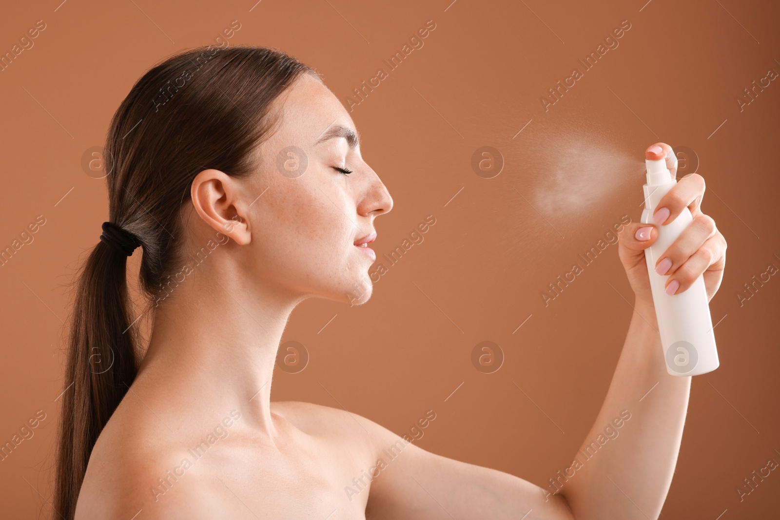 Beautiful woman spraying thermal water onto face on light brown background Photo of Beautiful woman spraying thermal water onto face on light brown background