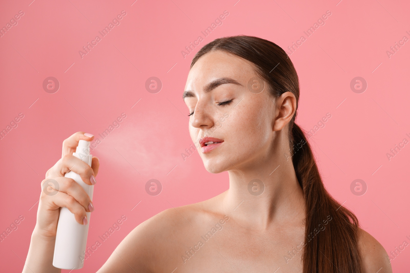 Beautiful woman spraying thermal water onto her face on pink background Photo of Beautiful woman spraying thermal water onto her face on pink background