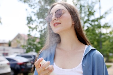 Photo of Smiling woman in sunglasses spraying thermal water onto her face outdoors