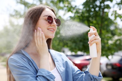 Photo of Smiling woman in sunglasses spraying thermal water onto her face outdoors