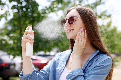 Photo of Smiling woman in sunglasses spraying thermal water onto her face outdoors