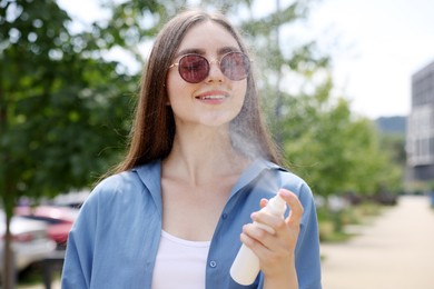 Photo of Smiling woman in sunglasses spraying thermal water onto her face outdoors
