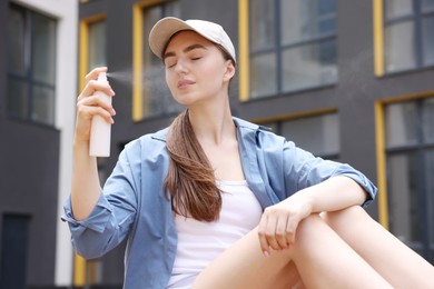 Photo of Beautiful woman in baseball hat spraying thermal water onto her face outdoors