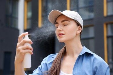 Photo of Beautiful woman in baseball hat spraying thermal water onto her face outdoors