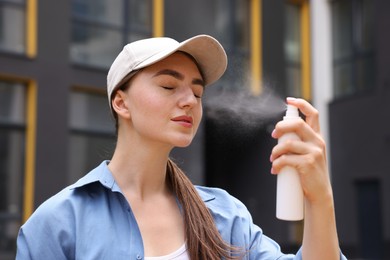 Photo of Beautiful woman in baseball hat spraying thermal water onto her face outdoors