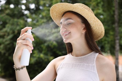 Photo of Smiling woman in straw hat spraying thermal water onto her face outdoors