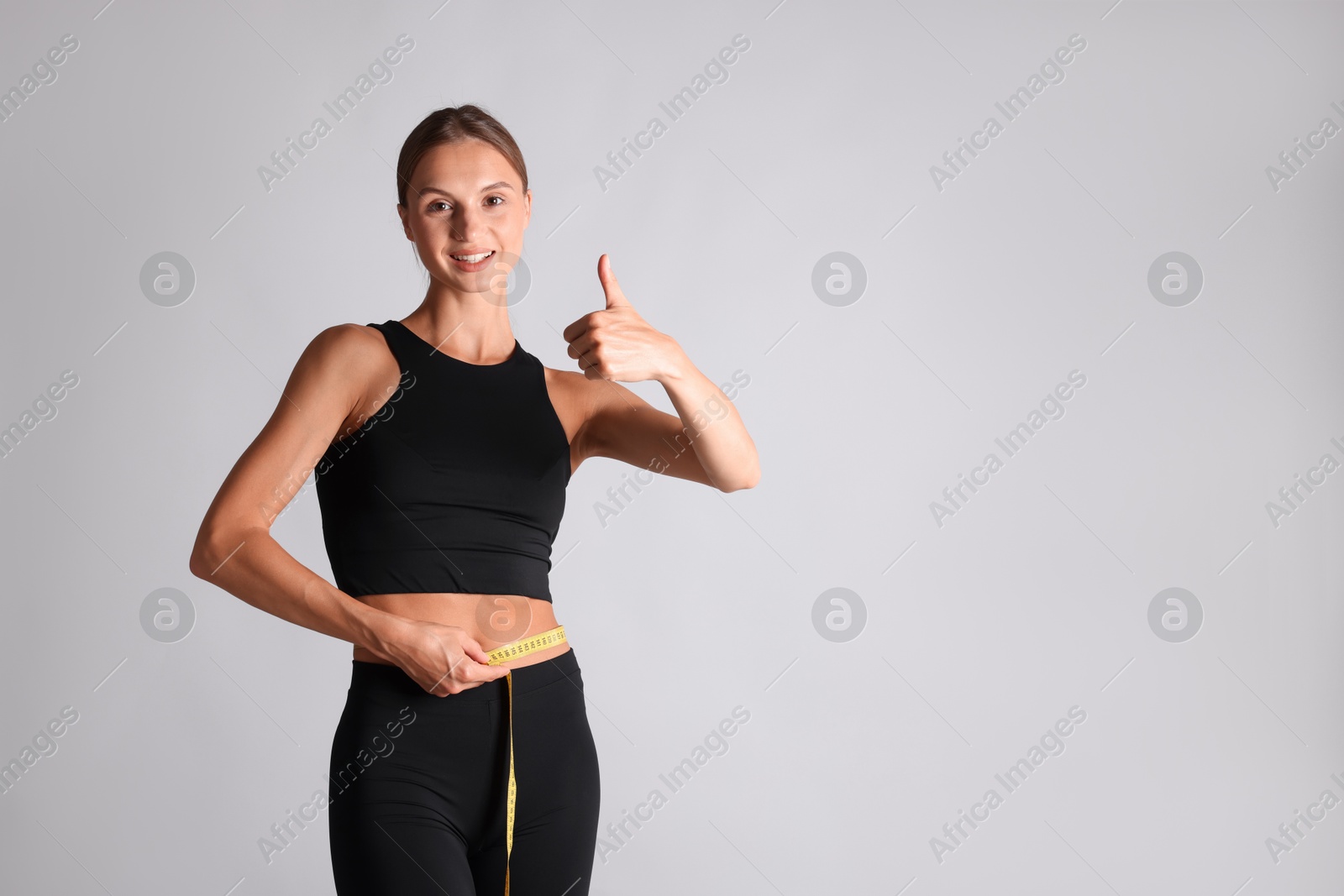 Woman measuring her belly with tape and showing thumbs up on light grey background, space for text Photo of Woman measuring her belly with tape and showing thumbs up on light grey background, space for text