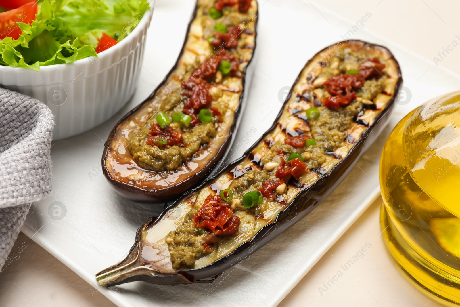 Photo of Slices of grilled eggplant with sun dried tomatoes, pesto and salad on white table, closeup