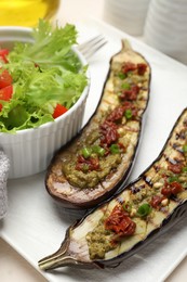 Photo of Slices of grilled eggplant with sun dried tomatoes, pesto and salad on white table, closeup