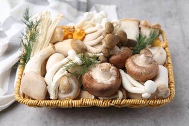 Photo of Different raw mushrooms and dill in wicker basket on grey textured table, closeup