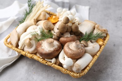 Photo of Different raw mushrooms and dill in wicker basket on grey textured table, closeup