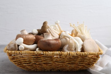 Photo of Different raw mushrooms in wicker basket on grey textured table against light background, closeup