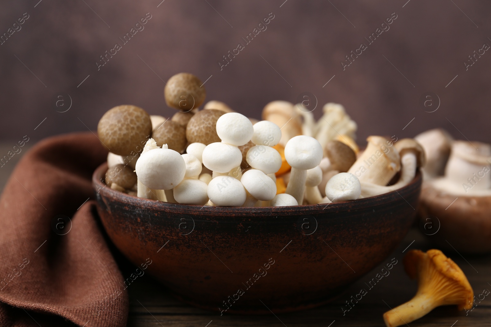 Different raw mushrooms in bowl on wooden table against brown background, closeup Photo of Different raw mushrooms in bowl on wooden table against brown background, closeup