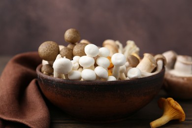 Photo of Different raw mushrooms in bowl on wooden table against brown background, closeup